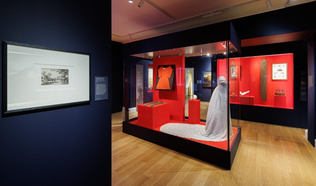 A photograph of a room in the museum show 'Untold Lives' at Kensington palace , where garments and work tools are on display in glass cases with red backdrops