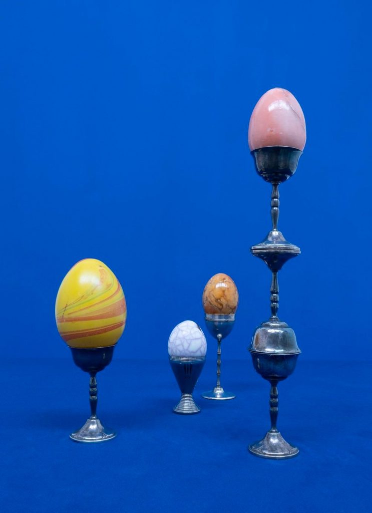 Photo of four different color crystal eggs in pewter candleholders of various heights against a bright indigo seamless studio backdrop.