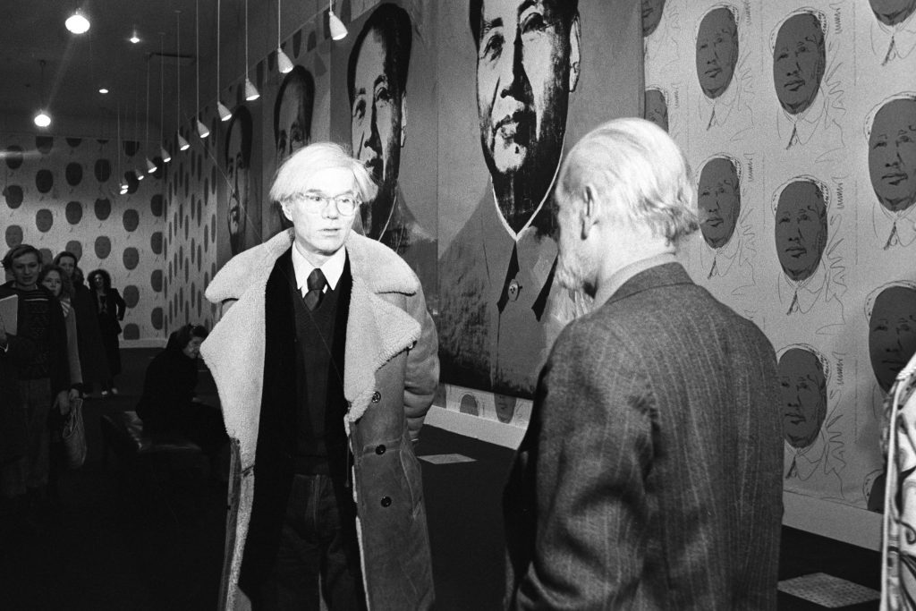 A man, artist Andy Warhol, standing in a gallery installed with his portraits of Mao Zedong.