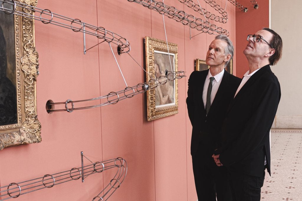 MAH Geneva Director Marc-Olivier Wahler (left) and artist Wim Delvoye (right) looking at Delvoye's artwork installed against a pink wall in one of the museum's gallery's.