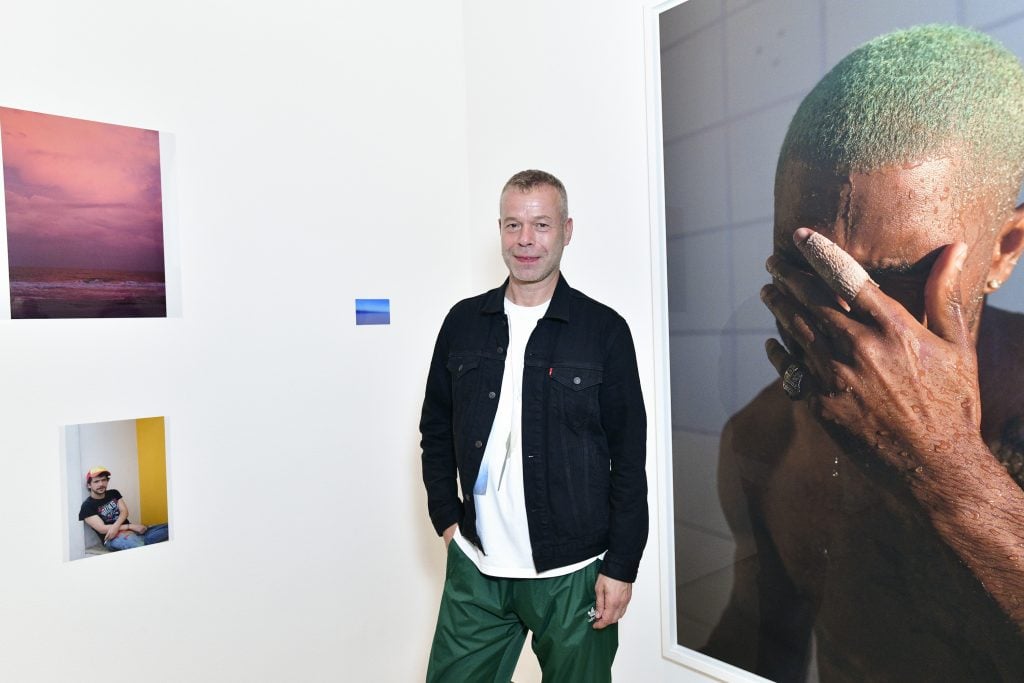Photographer Wolfgang Tillmans standing in a white-walled gallery hung with his photographs, including a somber portrait of rap star Frank Ocean.