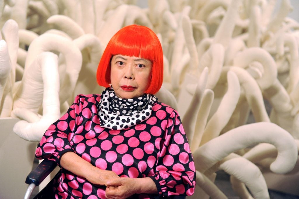 Artist Yayoi Kusama wears a bright red wig and pink polka dot dress in front of a white sculpture