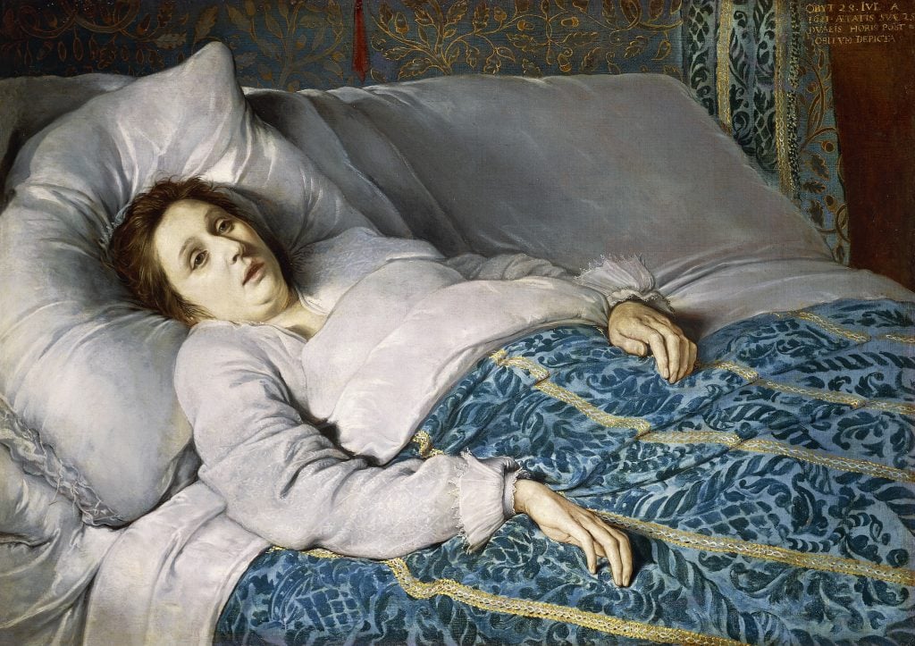 A pale woman lying in bed, a blank expression on her face.