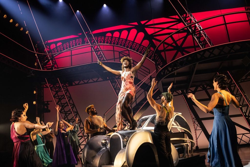 A performer exuberantly raises her arms center stage, standing on a vintage car, embodying a moment of triumph in a theatrical production. Surrounded by a cast dressed in colorful, 1920s-style costumes, the scene is set against a geometric, Art Deco-inspired backdrop bathed in warm and cool lighting. This dynamic tableau captures the essence of a vibrant musical set in a bygone era, filled with energy, passion, and the fashion of the time.