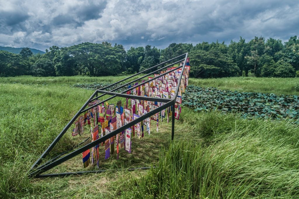 this picture shows a kite-like installation artwork with dozens of colourful embroidery pieces draped on the frame of the installation, mounted on a large piece of green grass land with lots trees at the back.
