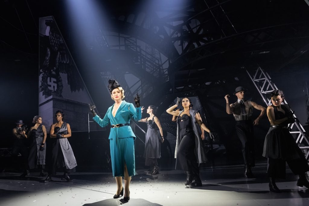 A woman in a striking turquoise suit with bold shoulders and black gloves commands the stage, flanked by dancers in dark, 1930s-inspired attire. Behind them, a monochrome set evokes the stark industrial lines of Art Deco design, interspersed with the ominous symbols of the Nazi regime, suggesting a narrative set in a tumultuous era. The scene captures the essence of a dramatic musical production, with the central figure exuding a sense of power and defiance amidst a tense historical backdrop.