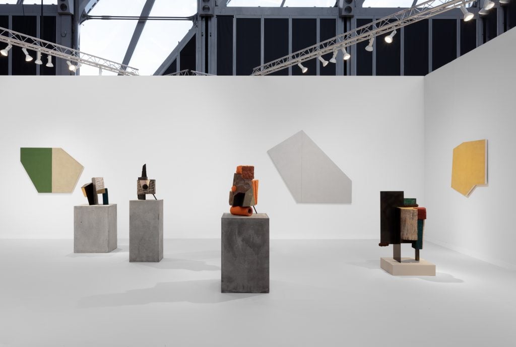 abstract geometric wood and steel sculptures are in a gallery surrounded by geometric minimal paintings