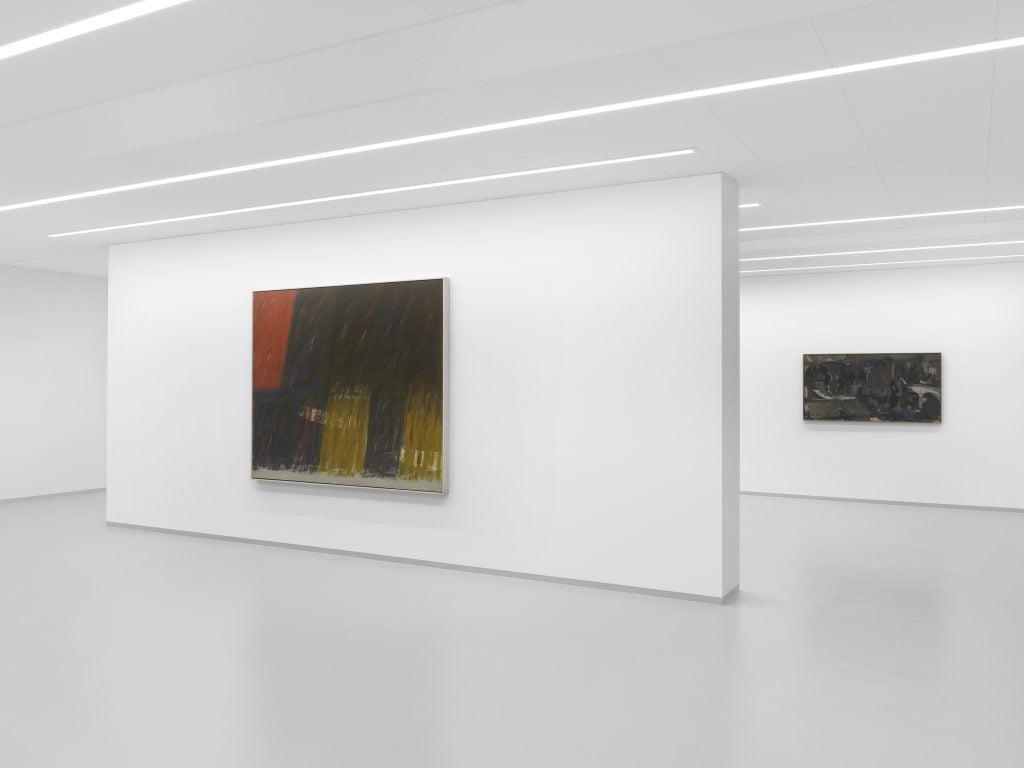 Two abstract paintings on fore and back wall of a white cube gallery space with three rows of lights along the ceiling.