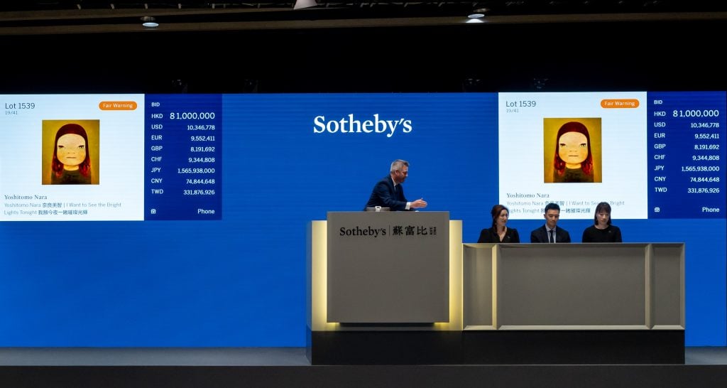 an auctioneer standing on the podium against a blue backdrop auctioning off a painting depicting the face of a girl