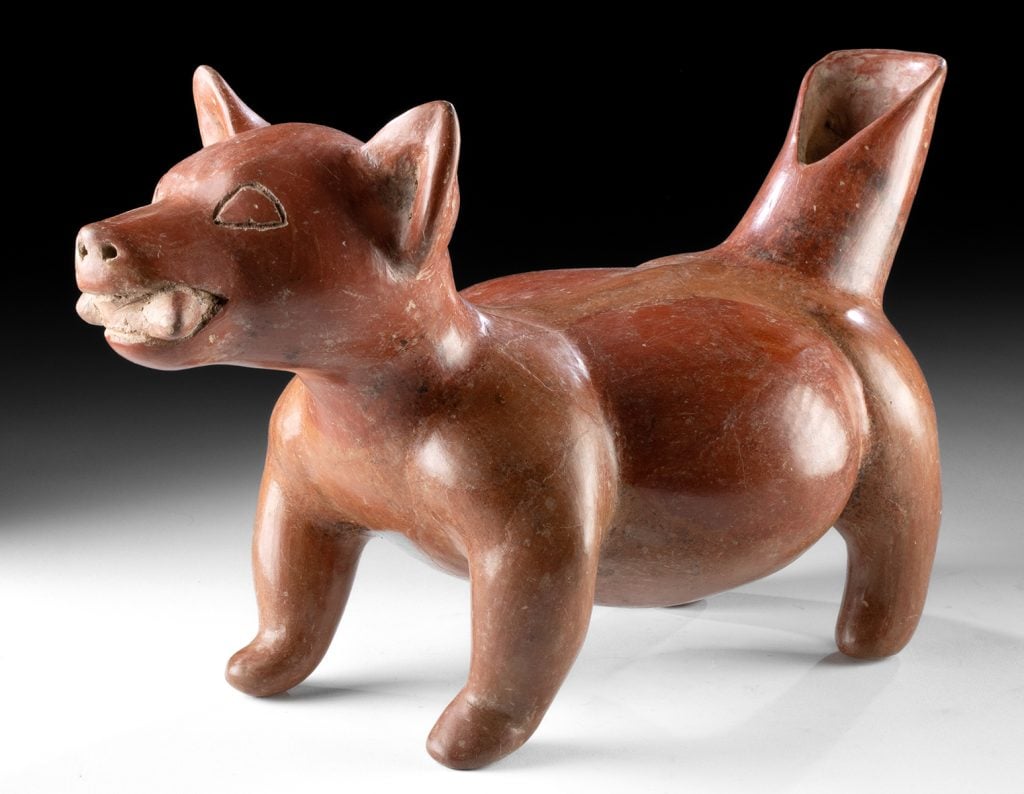 A ceramic sculpture of a dog with a reddish-brown glaze, depicting a hairless breed. This figure is likely representative of the Colima culture of ancient western Mexico, where such dog figures were commonly included in burials.