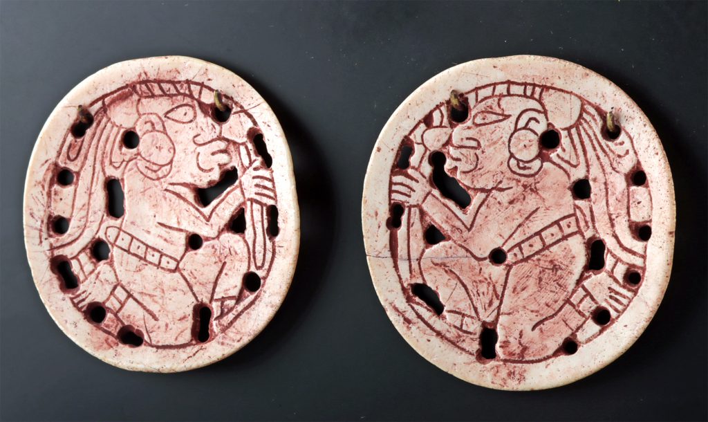 Two circular, pre-Columbian ceramic discs with red pictographs on a white background. Each disc has multiple holes around the perimeter and a central figure that appears to be playing a wind instrument. Their style suggests they may be from Mesoamerica, possibly Maya.