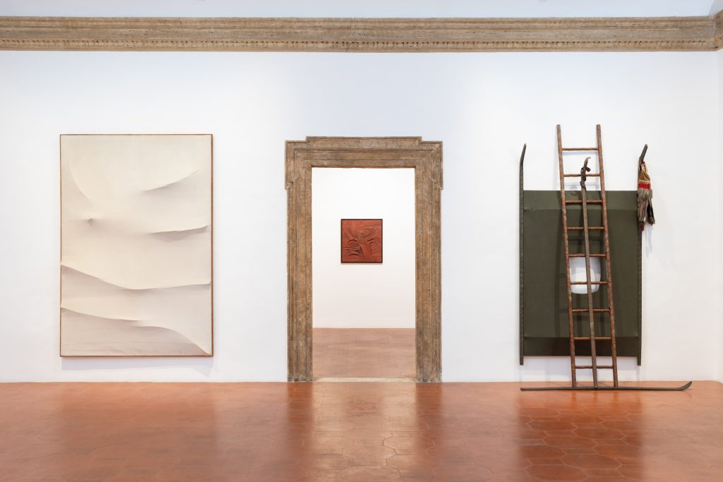 Installation view of works by salvatore scarpitta with a white monochrome painting on the left of a marble doorway with a structure under the canvas disrupting the flattness and an assemblage piece with a ladder on the right, and throuh the doorway a small red painting can be see on the far wall.