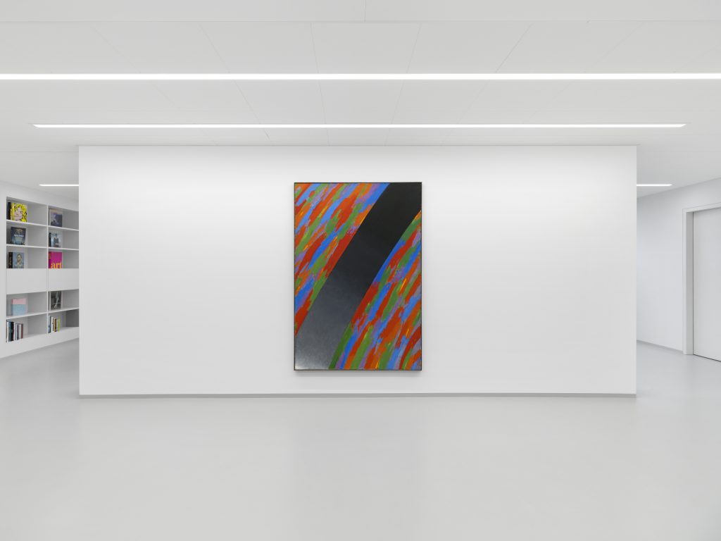 Abstract painting in a white cube gallery space, with the partial view of a bookshelf wall along the left side.