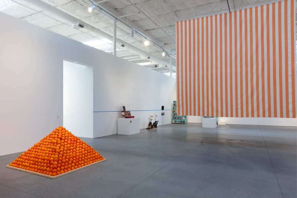 A color photo shows a capacious white-walled gallery with a sculpture of a pyramid of oranges and a painting with white and orange stripes