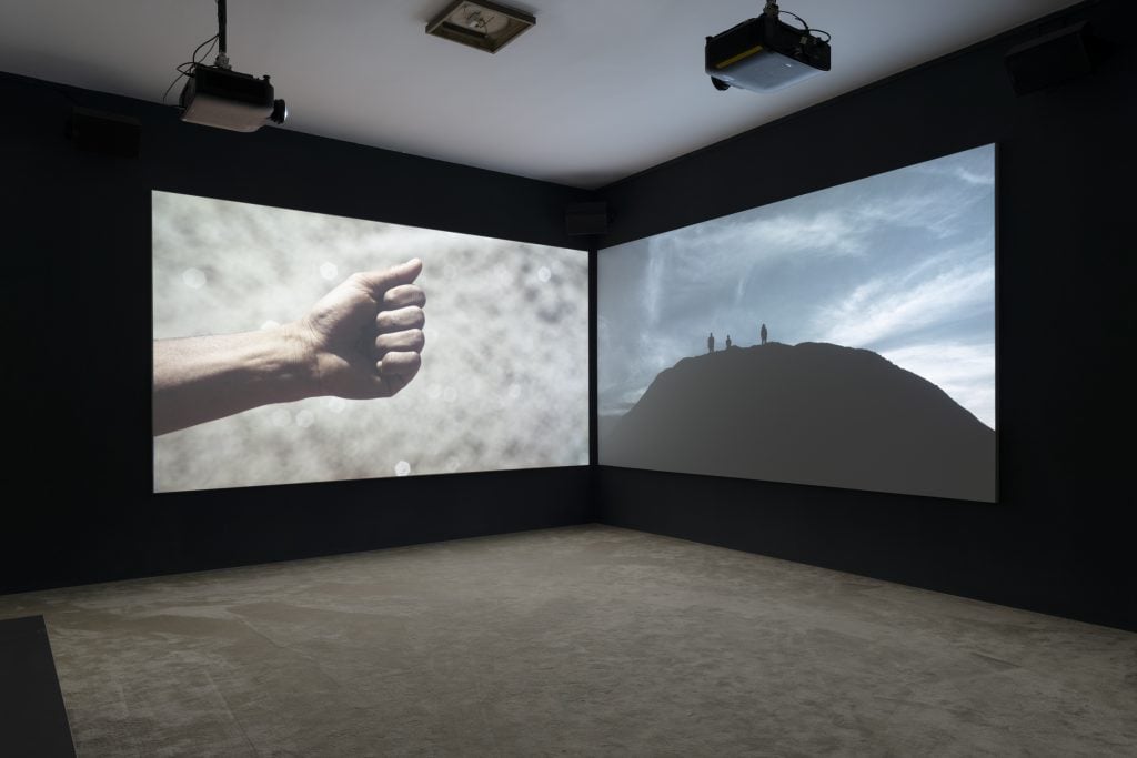 Two screens in a darkened room, one showing a closed fist and another the silhouette of a mound