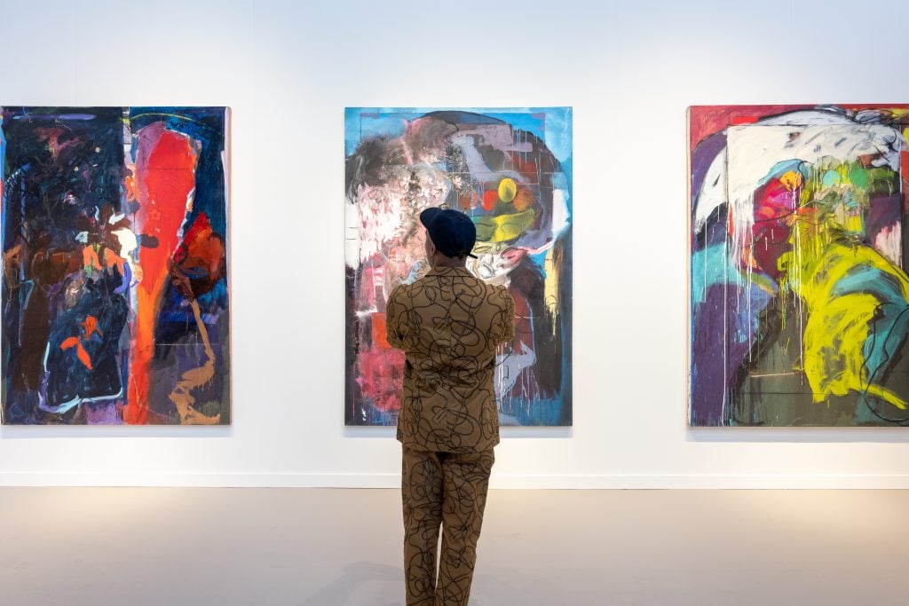 A man in a dark green suit with a swirly pattern, and a black baseball cap, stands with his back to us looking at three colourful abstract paintings on a white wall.