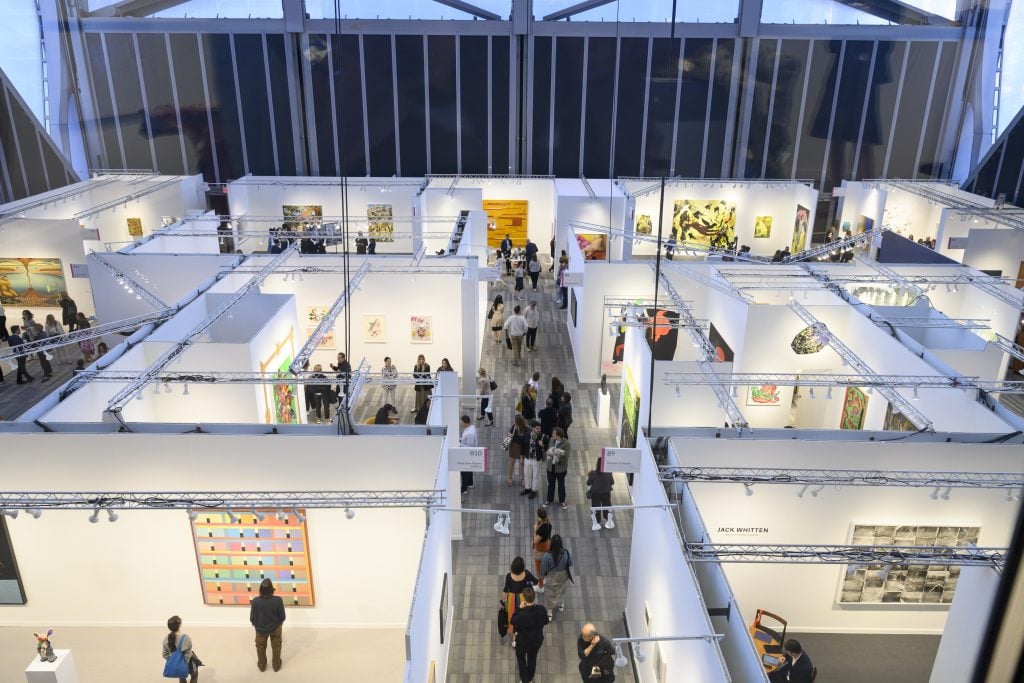 Brightly lit booths in an exhibition space show colourful paintings, the scene is photographed from above. 