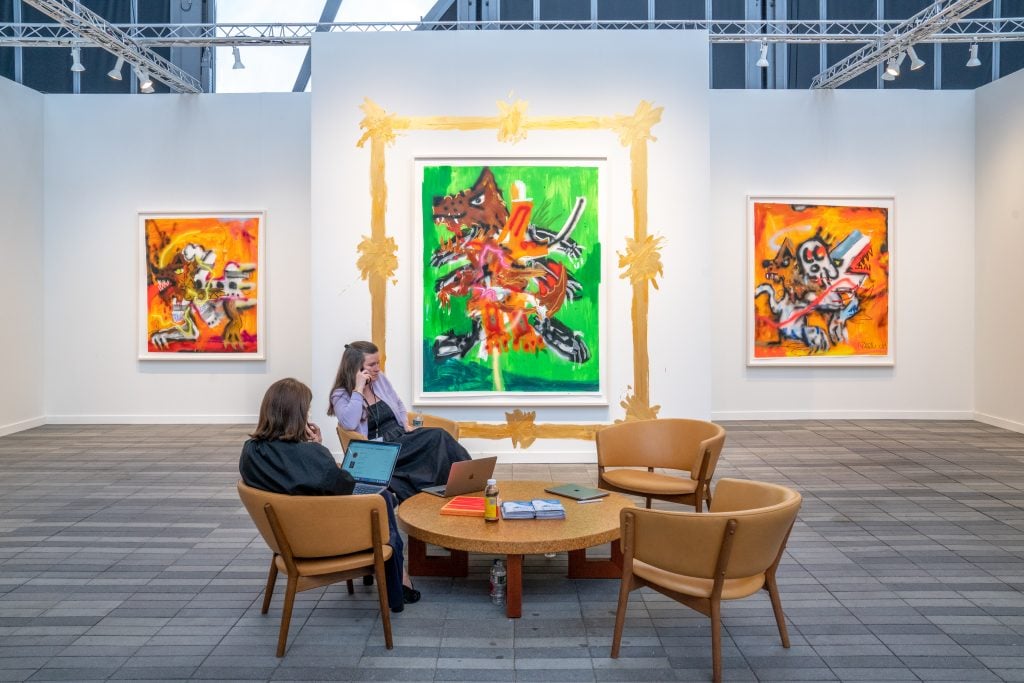 two women sit at a wooden table with colorful abstract paintings on the wall behind them