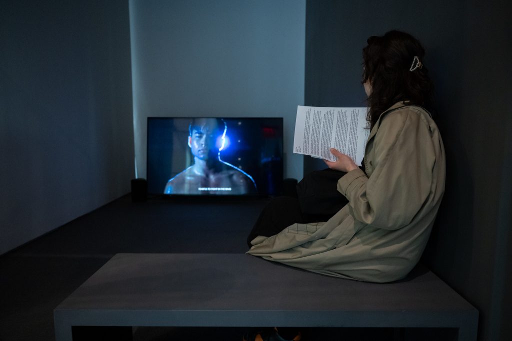a woman sits in a dark cubicle watching video art on a screen