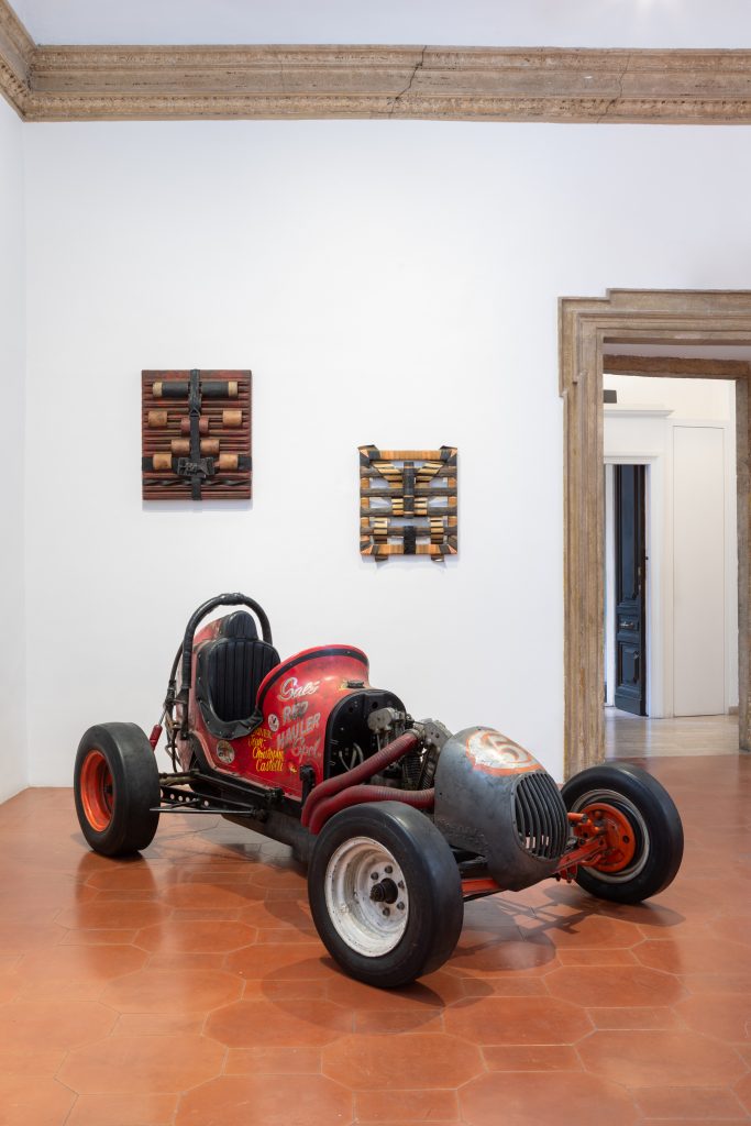 A dune buggy made of various parts sits in a gallery space with two small-scale paintings staggered on the wall behind it.