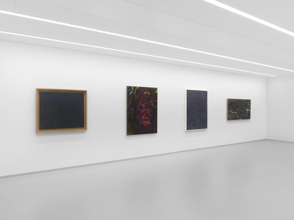 Four abstract paintings on the wall of a white cube gallery space with three rows of lights along the ceiling.