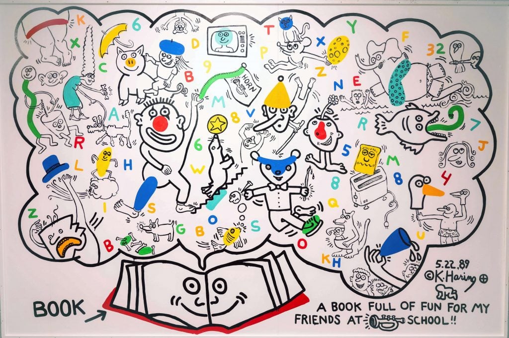 A vividly playful mural by Keith Haring presents a dynamic assortment of his signature animated characters and symbols, interspersed with letters and numbers, swirling around a central, grinning book. The artwork, titled "A Book Full of Fun for My Friends at School," is dated 5.22.89, capturing Haring's iconic style with its energetic lines and bright, primary colors. This engaging piece is an homage to literacy and learning, designed to inspire and delight young minds.