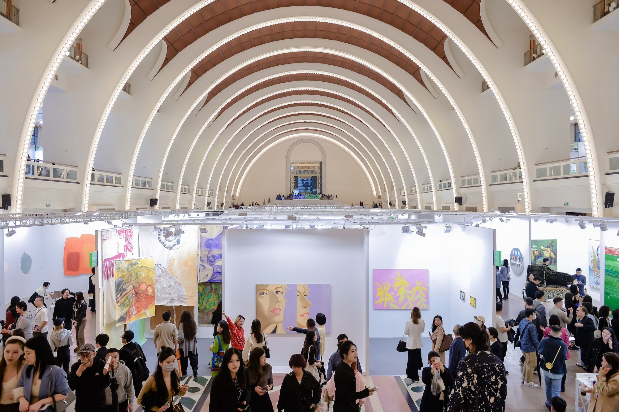 The image is of a large indoor hall with an arched ceiling. Crowds fill the hall.