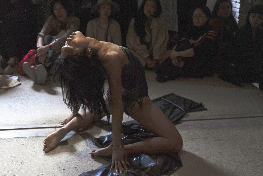 a queer performer dressed in a grey outfit kneeling on the ground during a performance