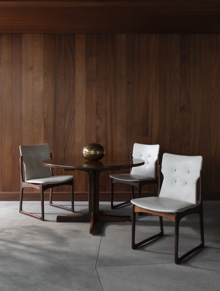 A danish design round dining table with four teak chairs with white fabric seats and backrests in a room with a gray floor and teak wood panelling.