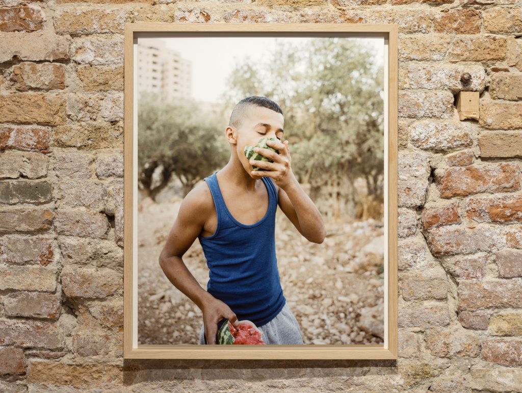 a photo of a young boy wearing a blue tank top and eating a watermelon hanging on a wall of exposed brick