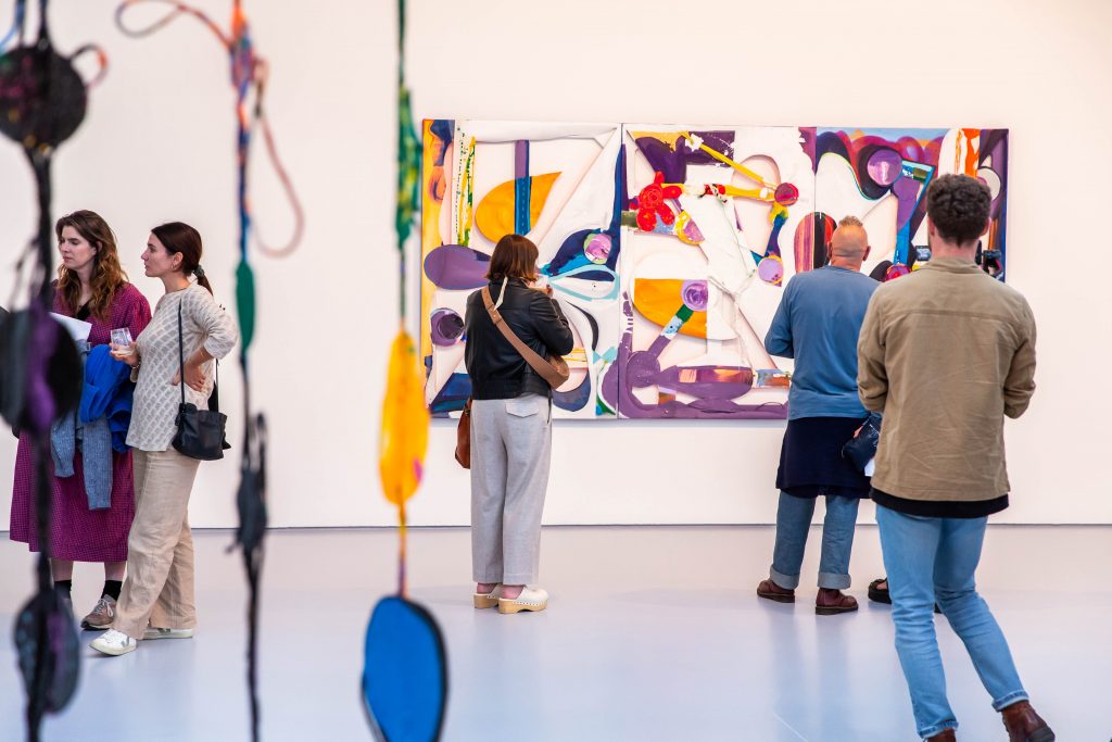 Several people stand In a gallery looking at a colourful painting and a blue and yellow sculpture