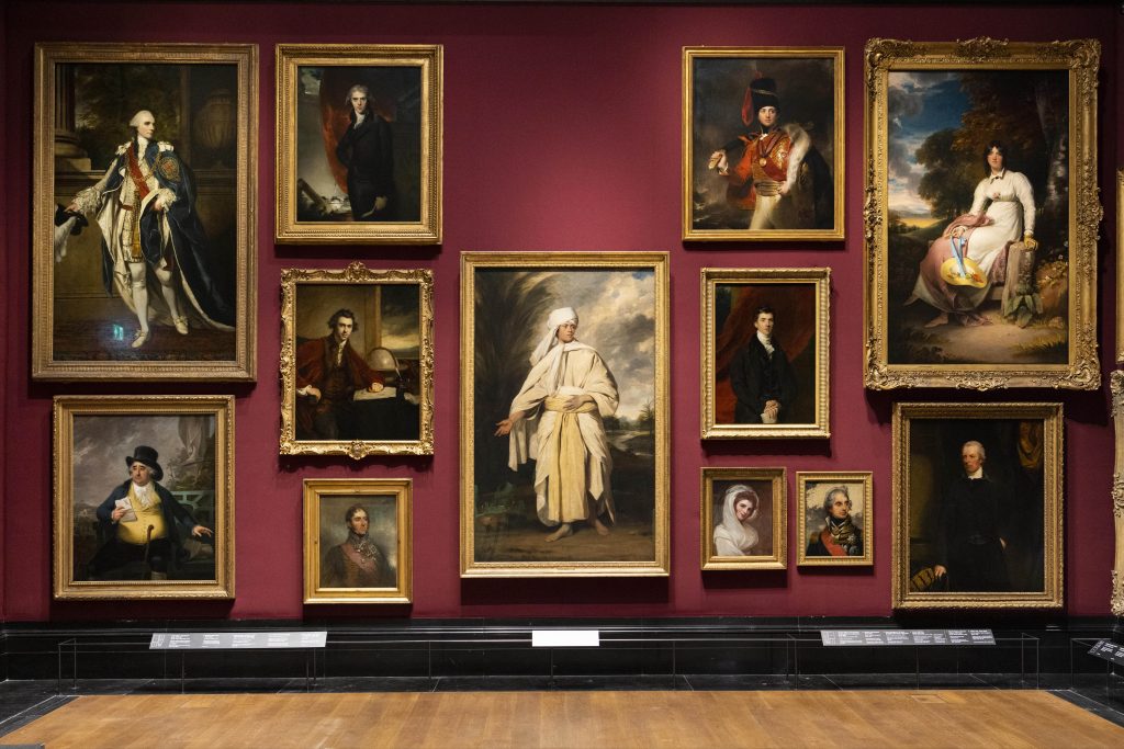 A gallery of paintings against a maroon wall in a museum.