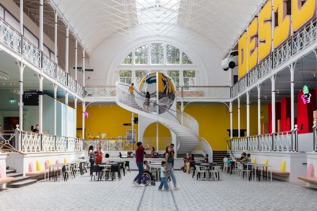 A large white gallery with a spiral staircase and a yellow painted wall. Visitors sit at chairs and tables.