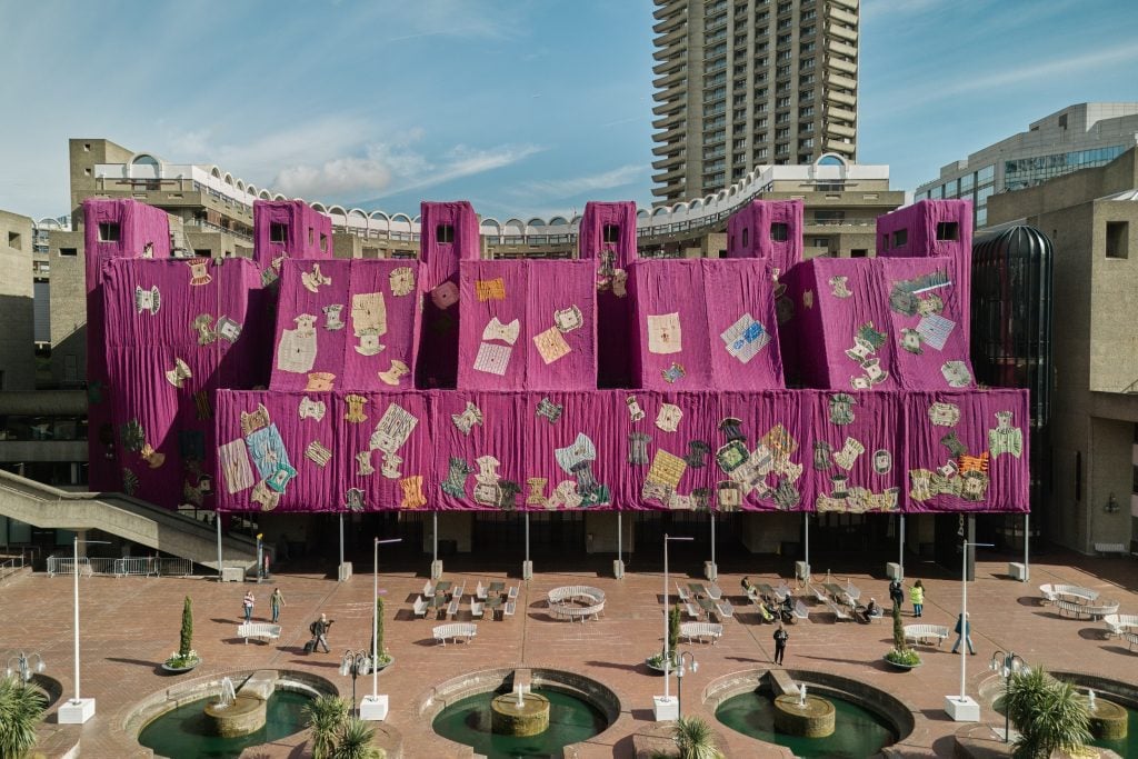 a front on view of a large bright pink tapestry draped like a tent over part of the Barbican building in London