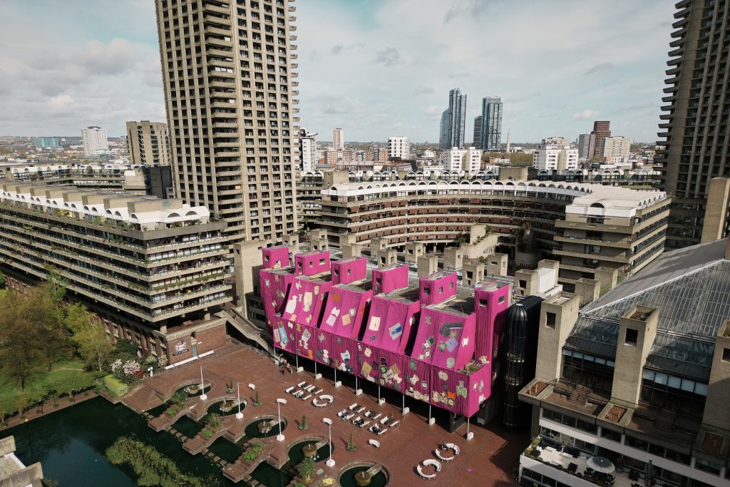 an aerial drone view of a large pink tapestry draped over part of the Barbican building in London