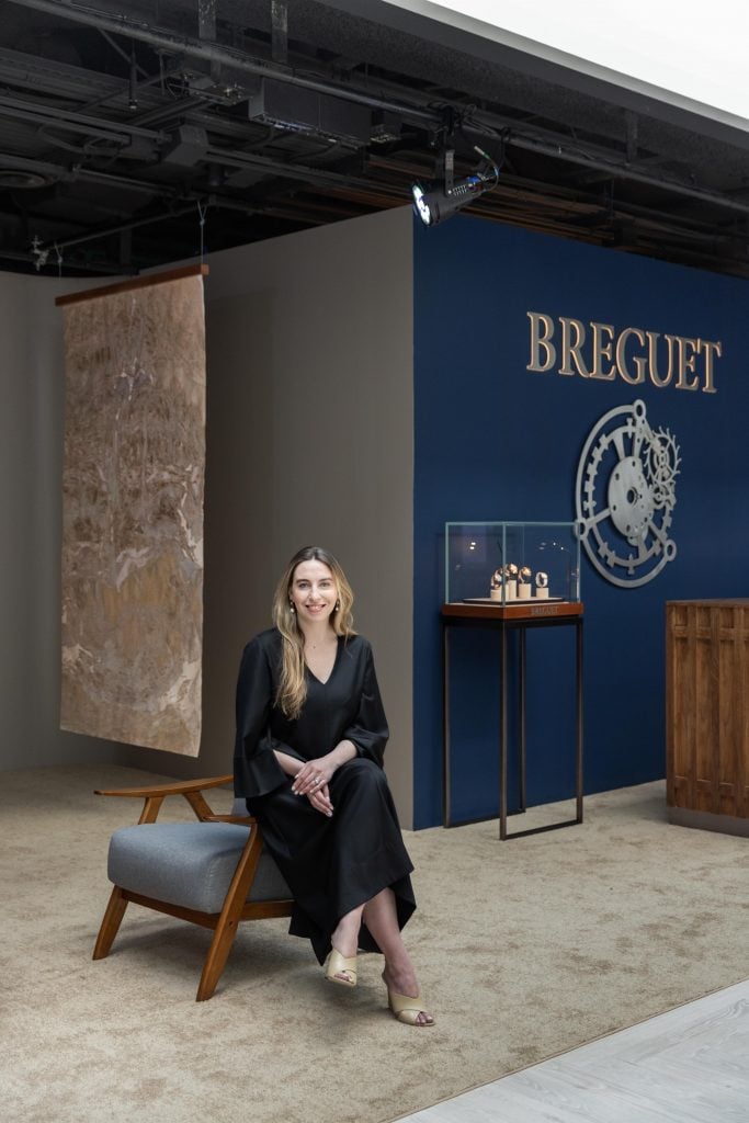 a Oman in a black dress sits atop a chair in front of an art installation by watch brand Breguet 