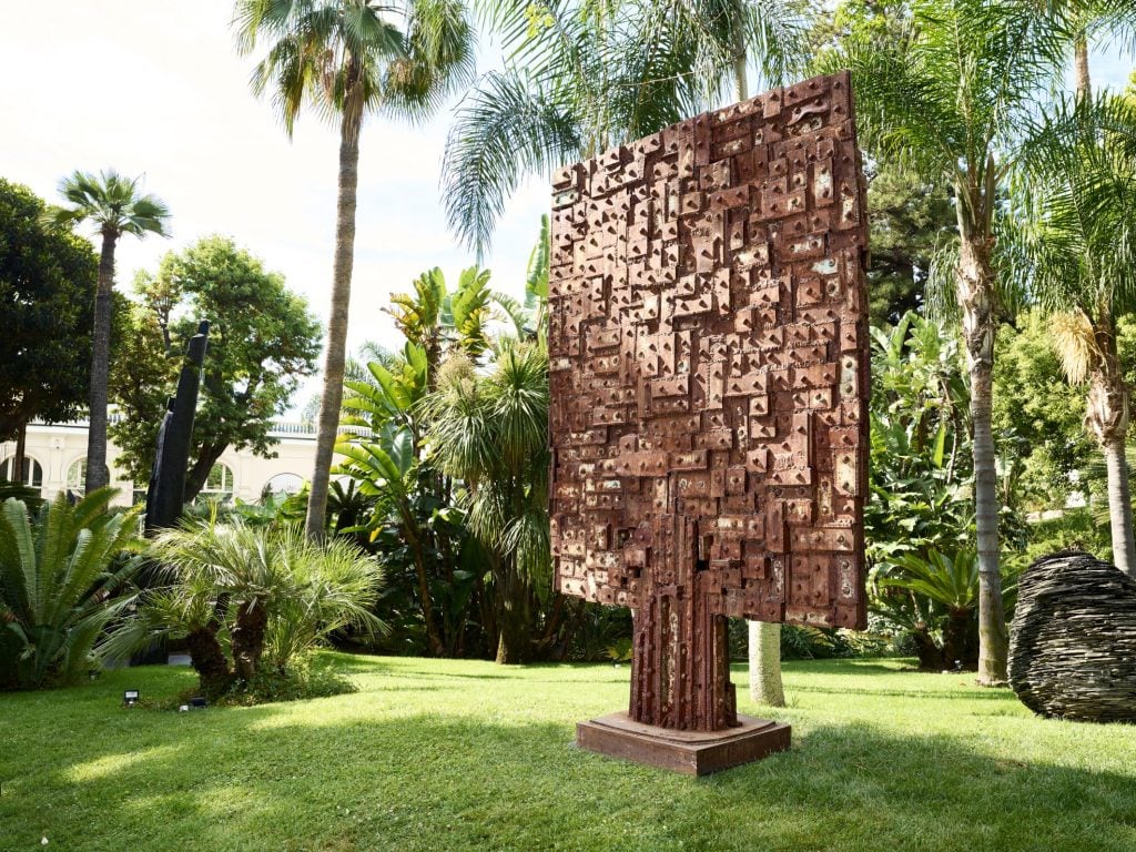 A rust red large-scale sculpture installed in a lush tropical landscape.