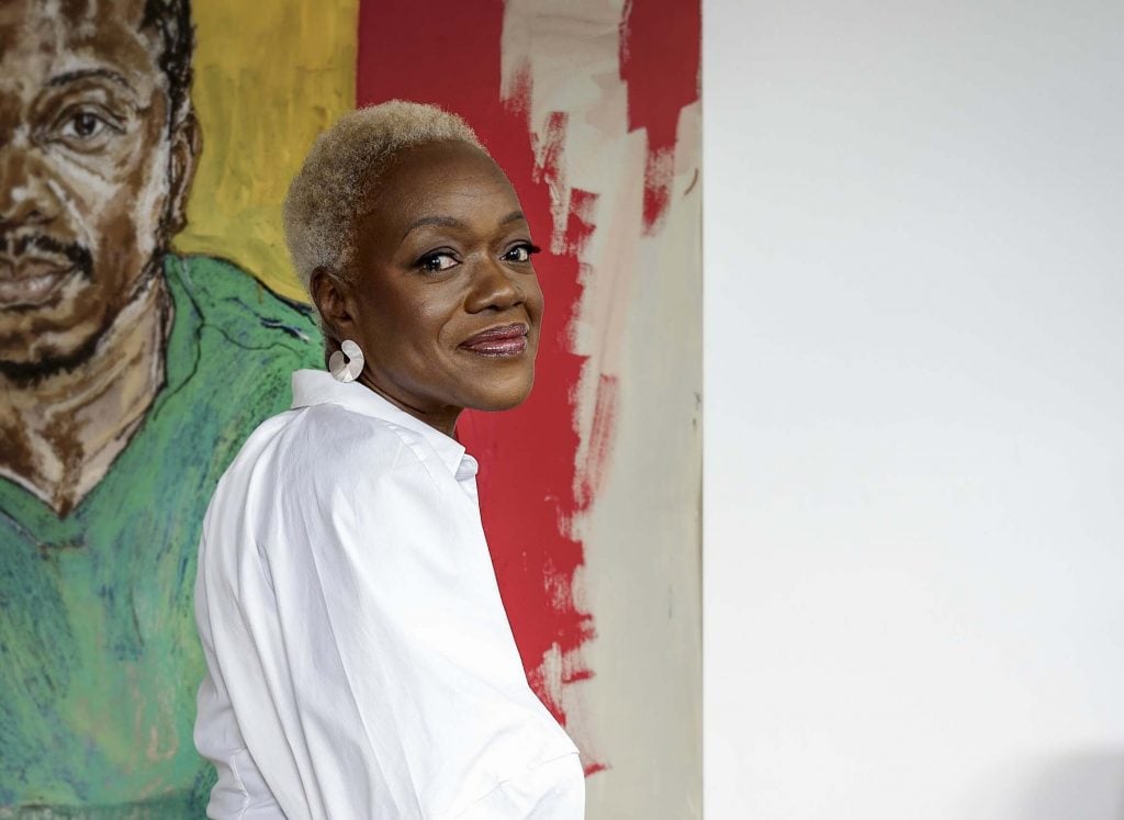a woman in a white shirt sits in front of a large painting of a black man in a green shirt. She is identified as the artist Claudette Johnson.