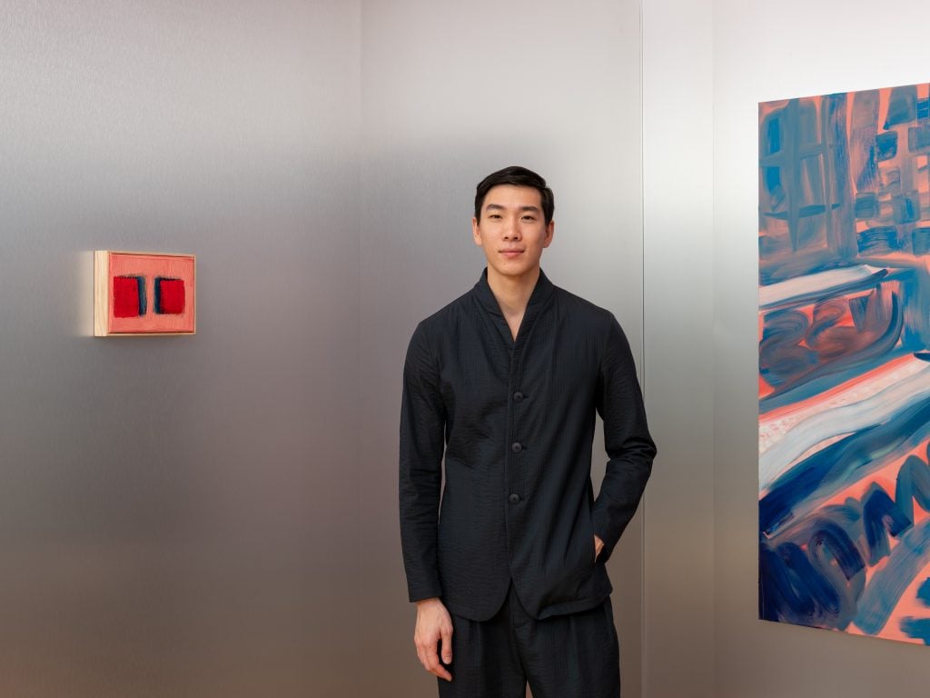 A 3/4 portrait of the artist wearing a black button down in a room with silver walls. A portion of an abstract painting is on view on the right, and a petite abstract painting about the size of a human hand in orange red and dark purple is on the wall on his right.