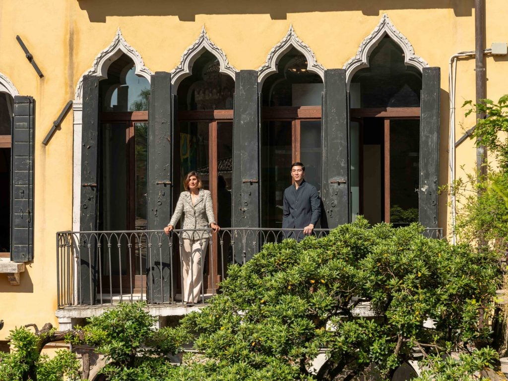 The curator and artist standing across a courtyard on a balcony surrounded by a row of windows with pointed cinquefoil windows.