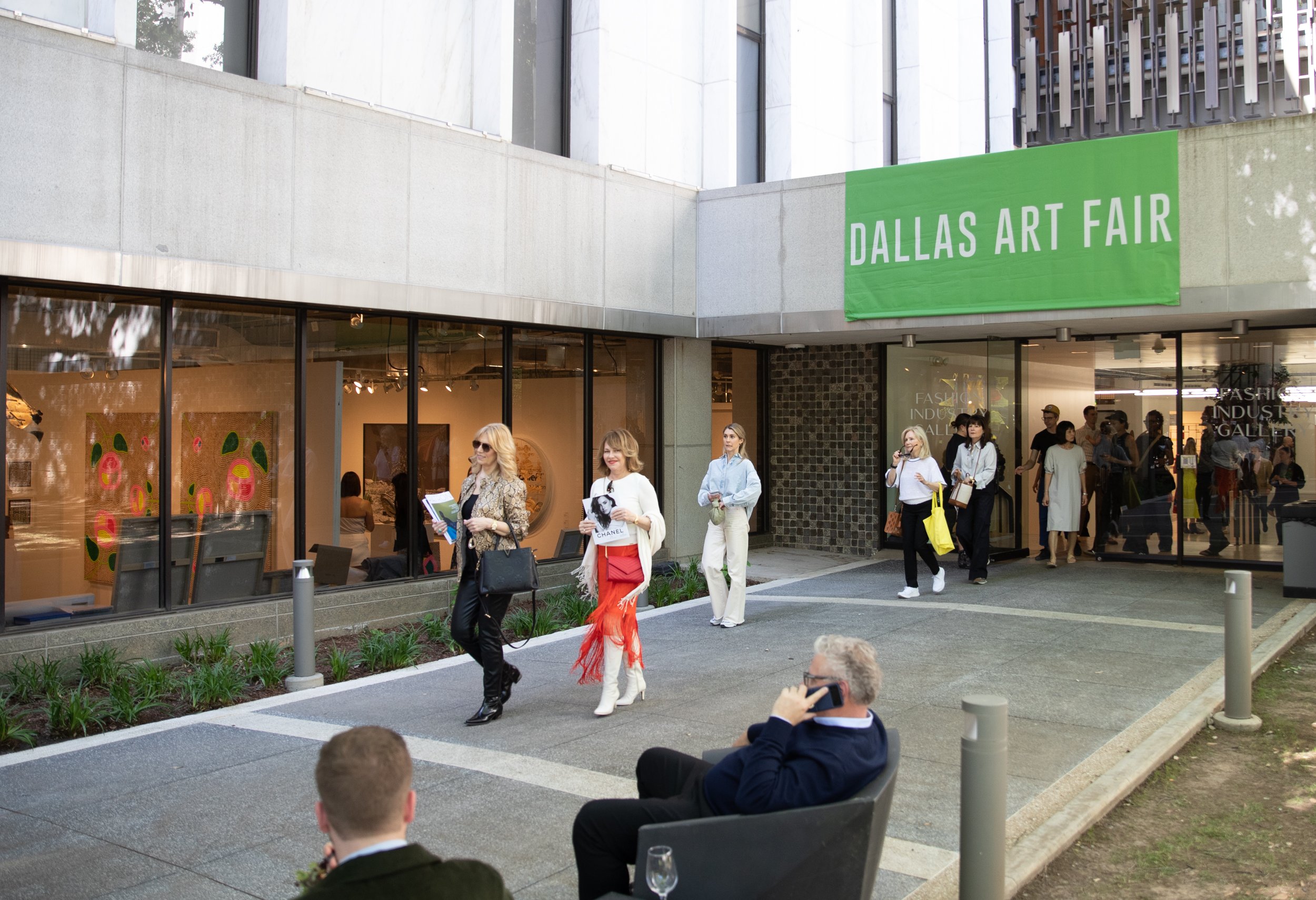 In a color photo, people walk outside on a walkway beneath a green sign that reads "Dallas Art Fair" in white letters.