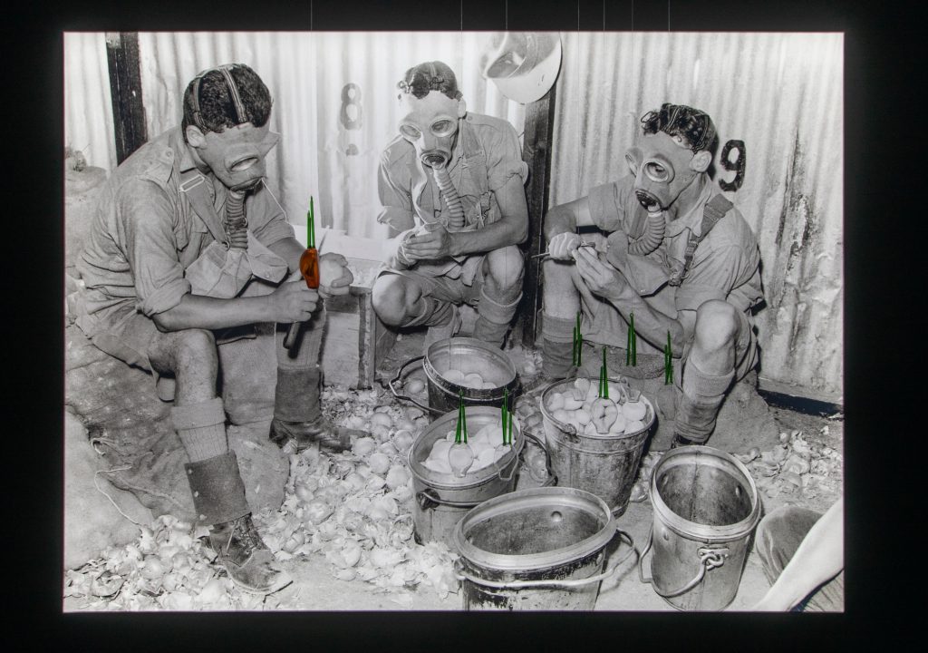 a black and white images shows three men wearing gas masks while peeling onions, there are some glass onions hanging in front of that image