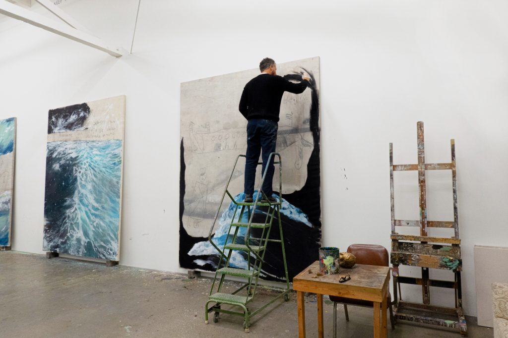 the artist working on a panting while standing on a ladder in his studio