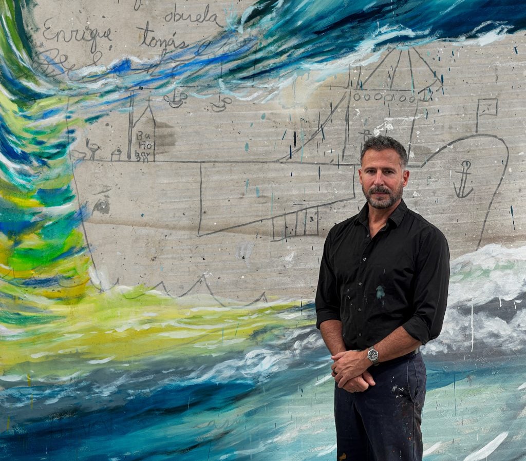 an image showing artist Enrique Martinez Celaya standing in front of his blue, green and white painting