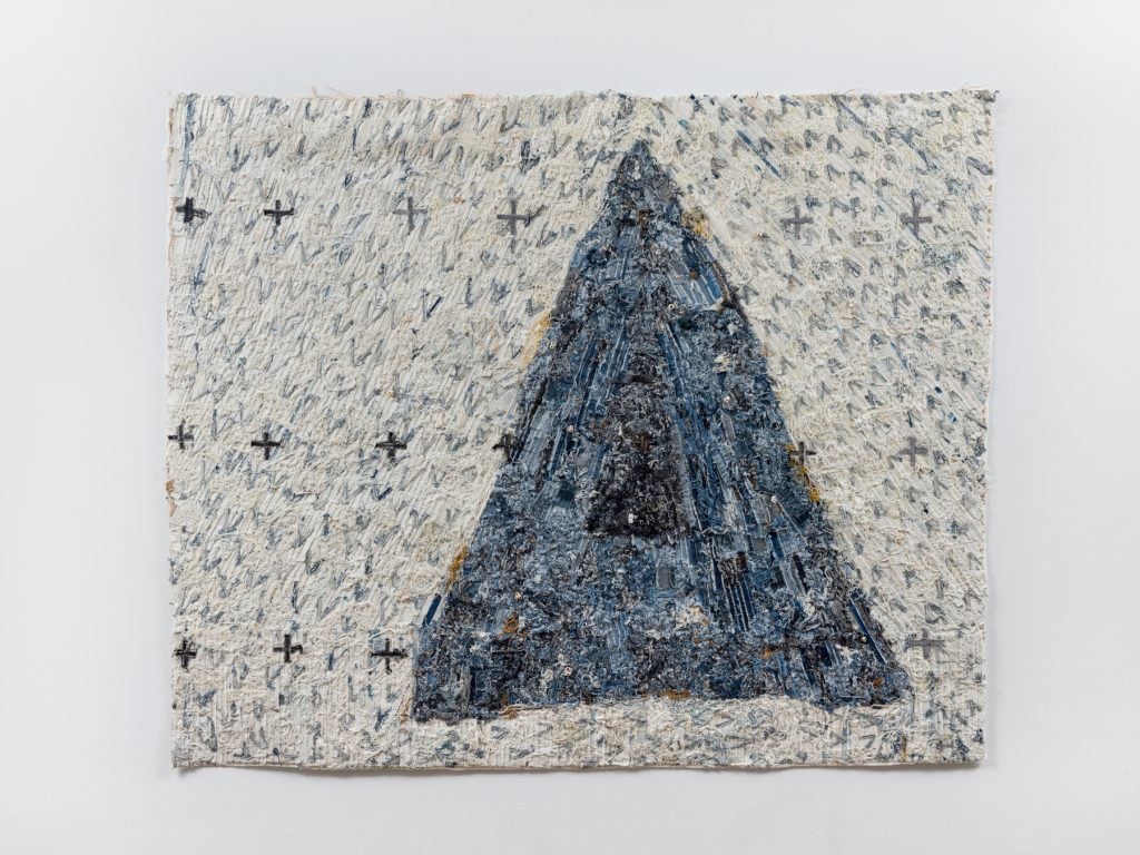 an image of an artwork by Jamal Cyrus feautring a blue pyramid on a white background made with bleached denim, denim, zippers, cotton, thread, sea shells, glue