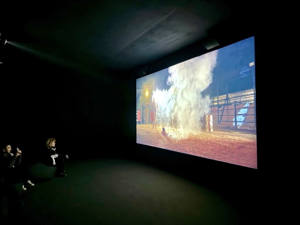 People in a darkened gallery watch a film where a ritual involving a cloud of smoke is happening