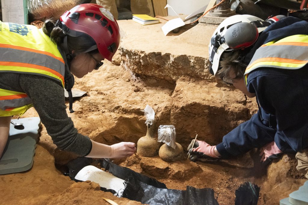 Two archaeologists dig around two glass bottles in the earth