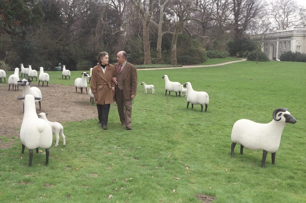 An older married couple walks through a green lawn filled with realistic sheep sculptures