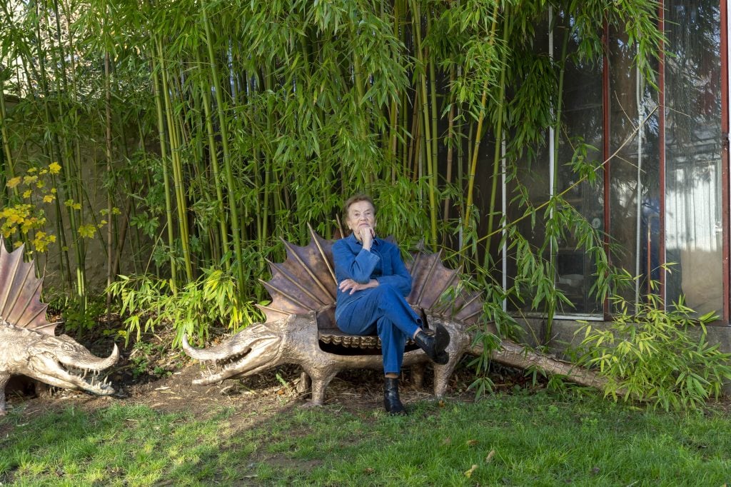 Artist Claude Lalanne sits atop a a bench that looks like a dinosaur in a backyard