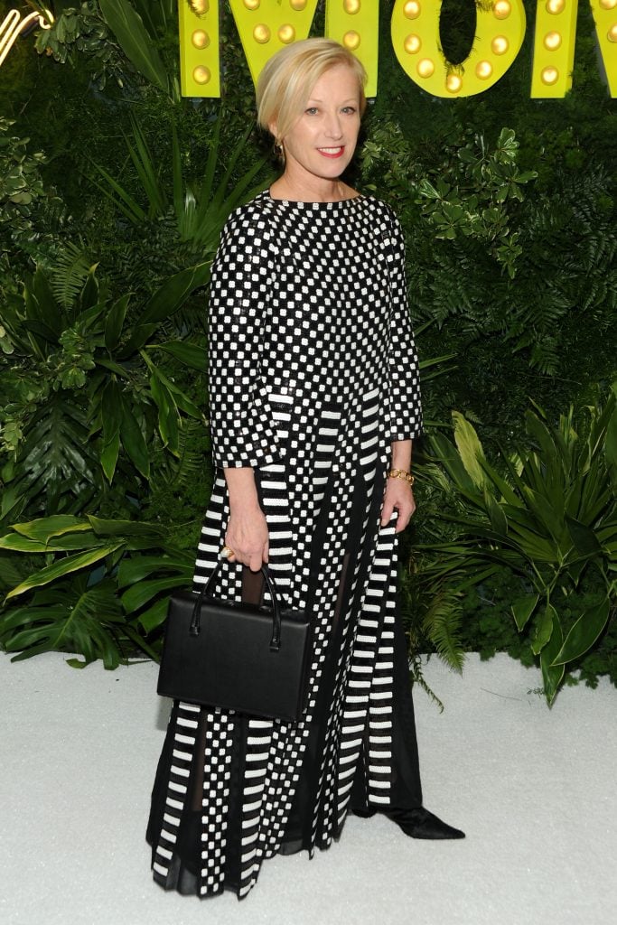 A full length, front fracing photo of Cindy Sherman wearing a black and white dress and holding a black purse before a leafy green background with yellow letters in it